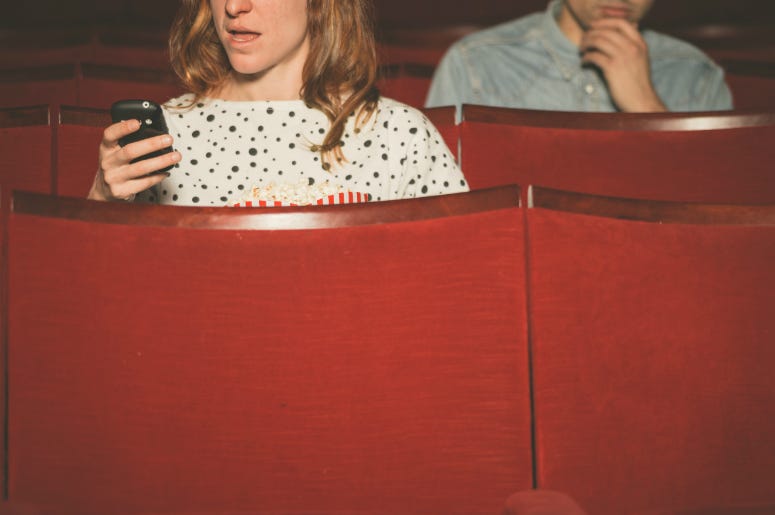 © Loganban | Dreamstime.com - <a href="https://www.dreamstime.com/stock-photo-woman-using-her-phone-theater-young-women-men-sitting-behind-image52737286#res17454685">Woman using her phone in theater</a> Woman using her phone in a theater as young man sitting behind her looks on.