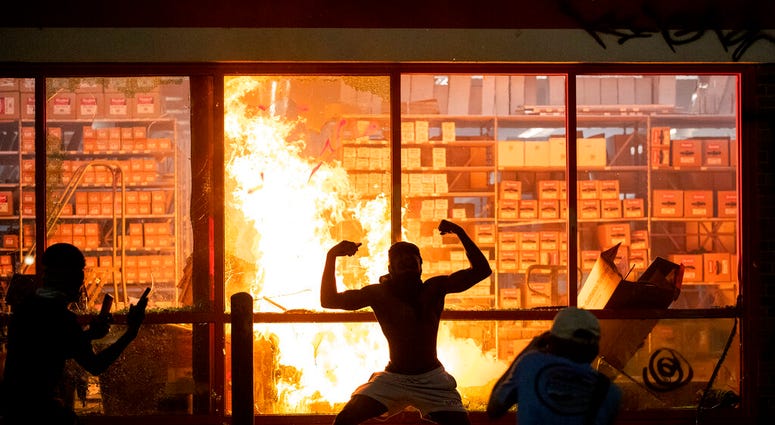 A man poses for photos in front of a fire at an AutoZone store, while protesters hold a rally for George Floyd in Minneapolis on Wednesday, May 27, 2020.