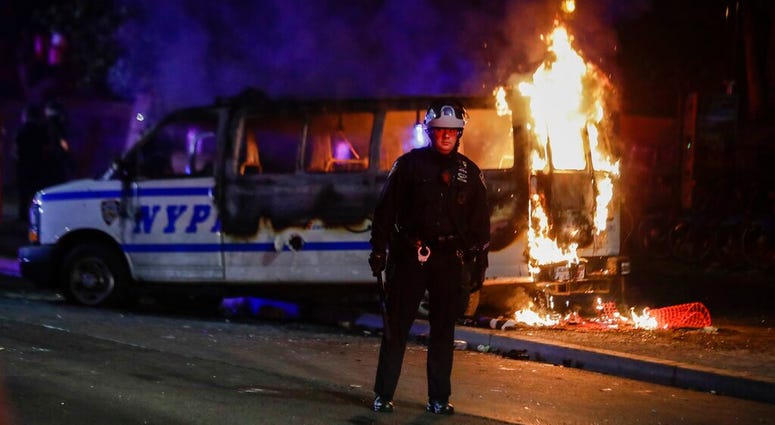A police officer watches a crowd as a police vehicle burns near Fort Greene Park in the Brooklyn borough of New York after protesters rallied outside Barclays Center over the death of George Floyd, a black man who died Memorial Day while in police custody