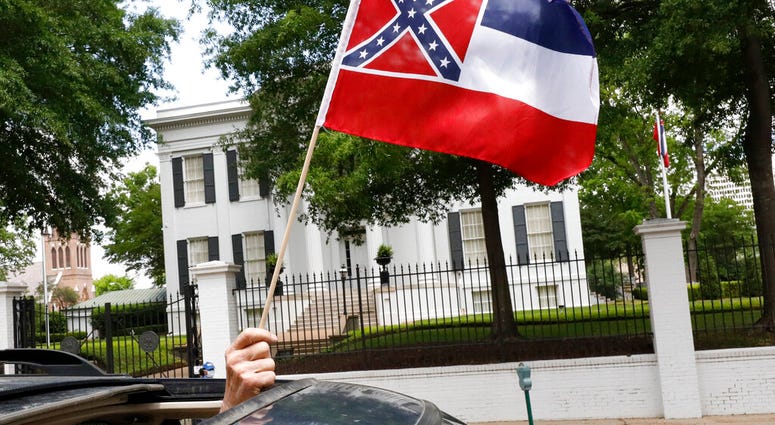 In this April 25, 2020 photograph, a small Mississippi state flag is held by a participant during a drive-by "re-open Mississippi" protest past the Governor's Mansion, in the background, in Jackson, Miss.