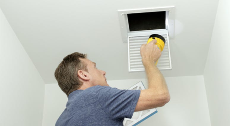 Man inspecting air ducts shining a flashlight through a small square ceiling vent into ducting pipes.