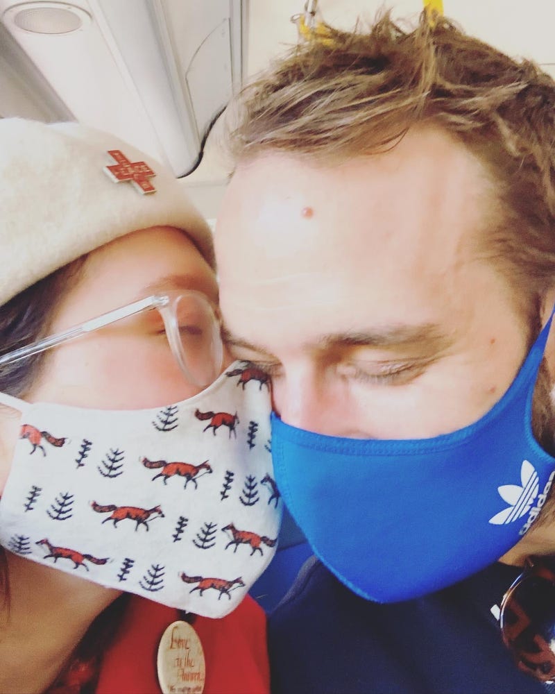 A woman kissing a man on the cheek, both are wearing masks