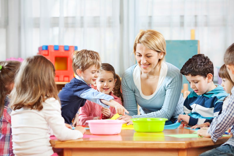 A baby sitter sits with several children around a table with paper and colored pencils