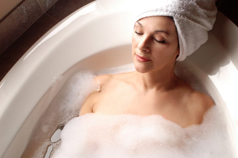 A woman with a towel wrapped around her head lies in a sudsy bath