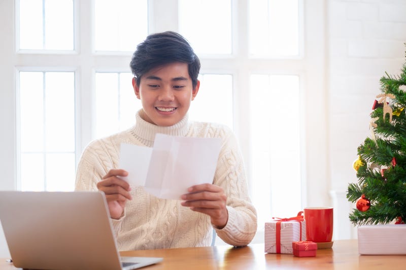 A man opens an envelope smiling at the card inside, with a Christmas tree and gifts nearby