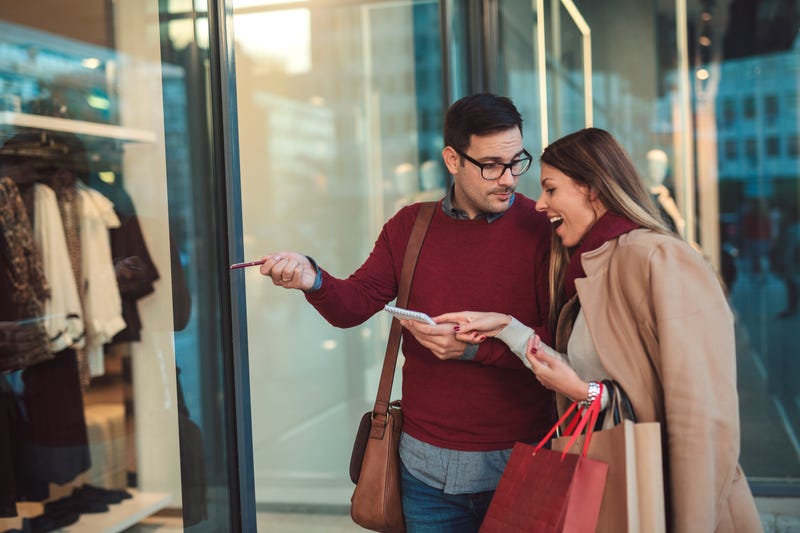 A man points to an item in a shop window while a woman points to the list in his hand