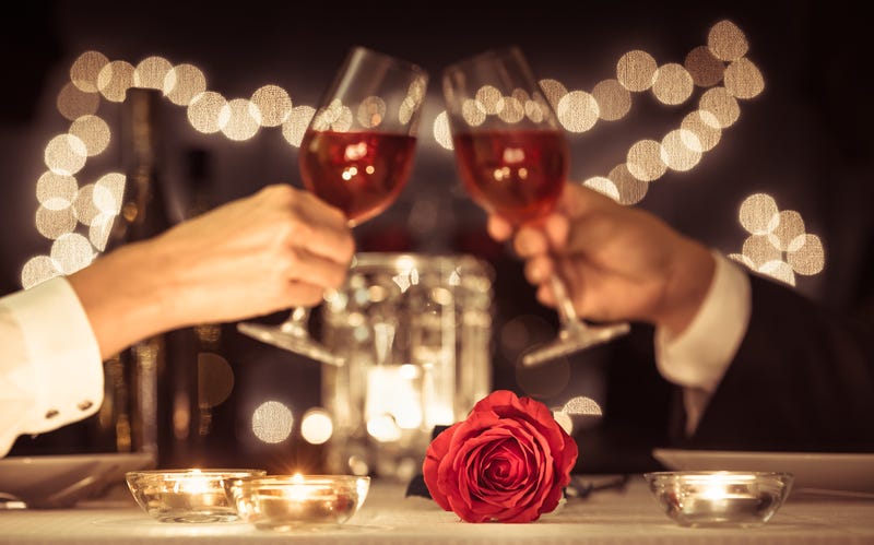 Two people toast with wine in the background of a table set with a rose and candles