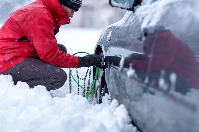 A man kneels down in the snow to apply snow chains to the tires of his car