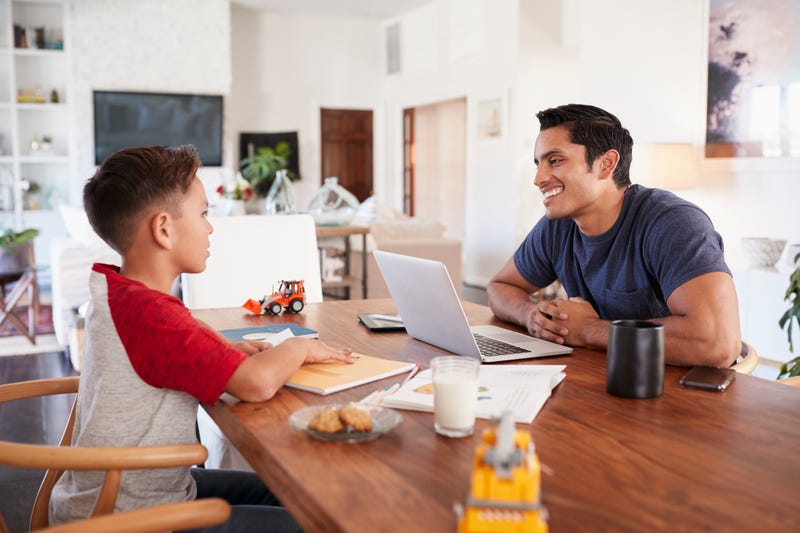 A man works at a laptop while his kid works across from him at a desk