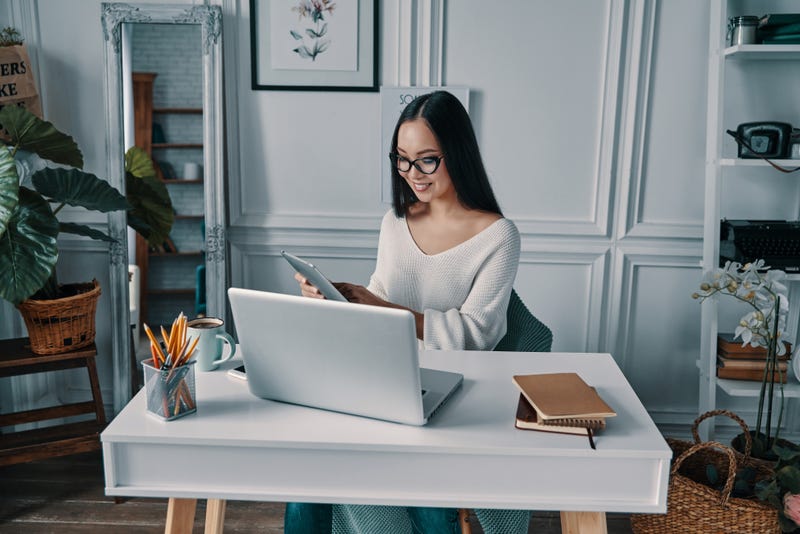 A woman in a home office sits at a desk with several devices and office supplies