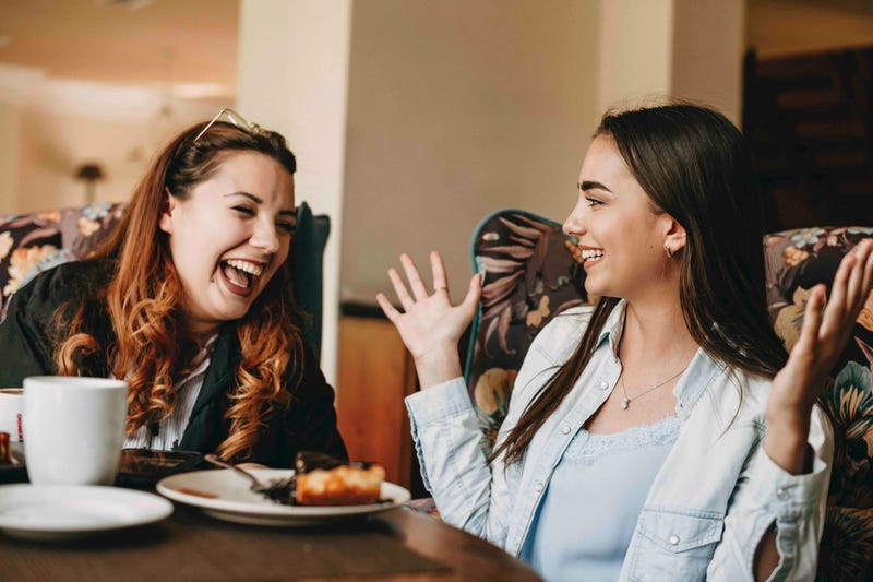Two women sit at a restaurant table, one making the other laugh with a story