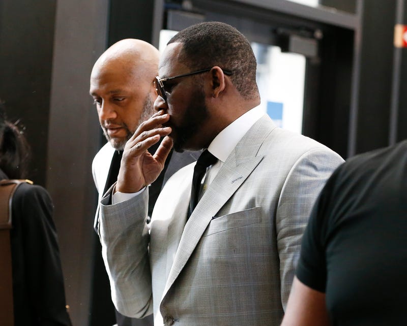 R. Kelly in a gray suit talks to a man next to him