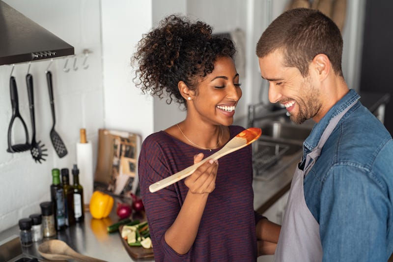 A woman feeds a taste of tomato sauce from a wooden spoon to a man in an apron in front of a kitchen counter where ingredients are displayed