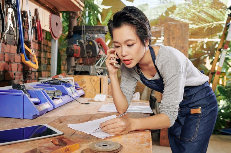 A woman on the phone leans over a work desk with pen and paper