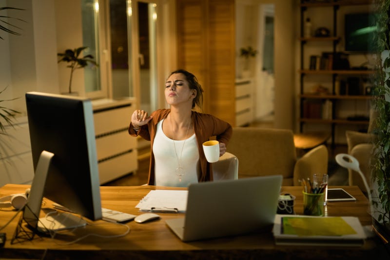 A woman sitting at a home computer stretches her back