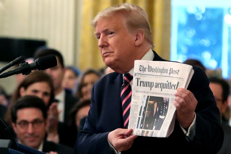 president donald trump holds up a newspaper reading with the headline 'trump acquitted'