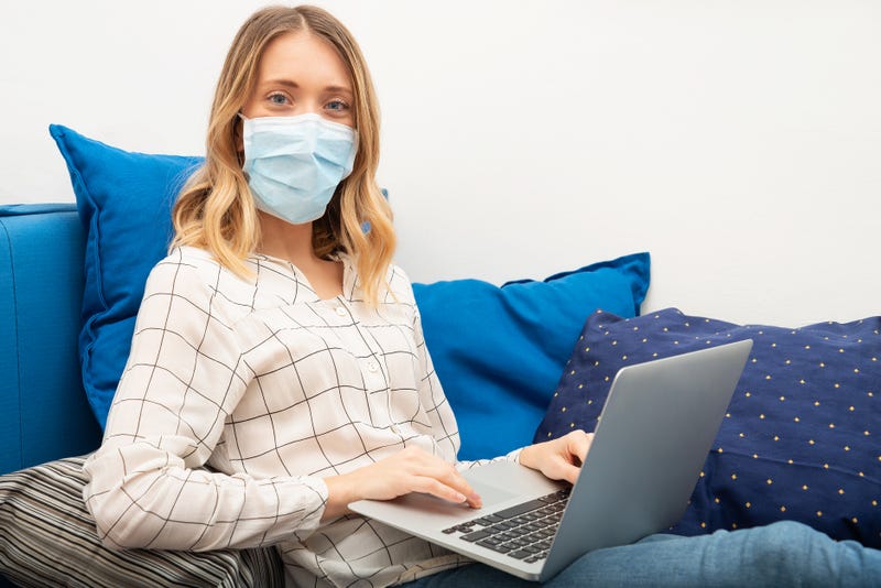 A woman in a medical face mask sits with a laptop on a couch