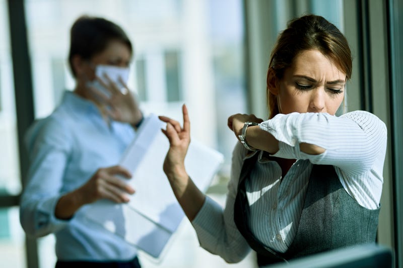 woman coughing into elbow with woman behind her in face mask