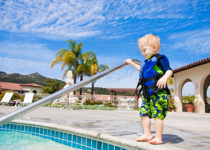 A cute little boy with a life jacket bravely steps into an outdoor swimming pool in San Diego