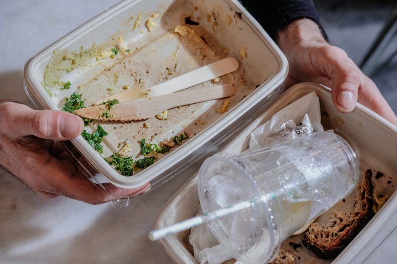 Man hands holding paper food containers, plastic cup, paper straw, wooden utensils to trash after meal, from above.