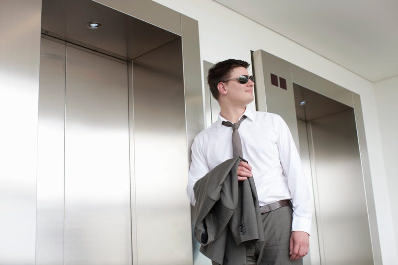 A man in sunglasses with his jacket over his arm waits for an office elevator