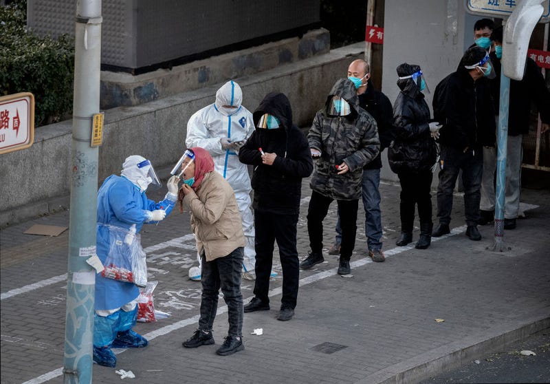 An epidemic control worker wears PPE to prevent the spread of COVID-19 as he gives a nucleic acid test to a resident as others wait in line wearing face shields before leaving by bus from an area with communities in lockdown on November 30, 2022 in Beijing, China.