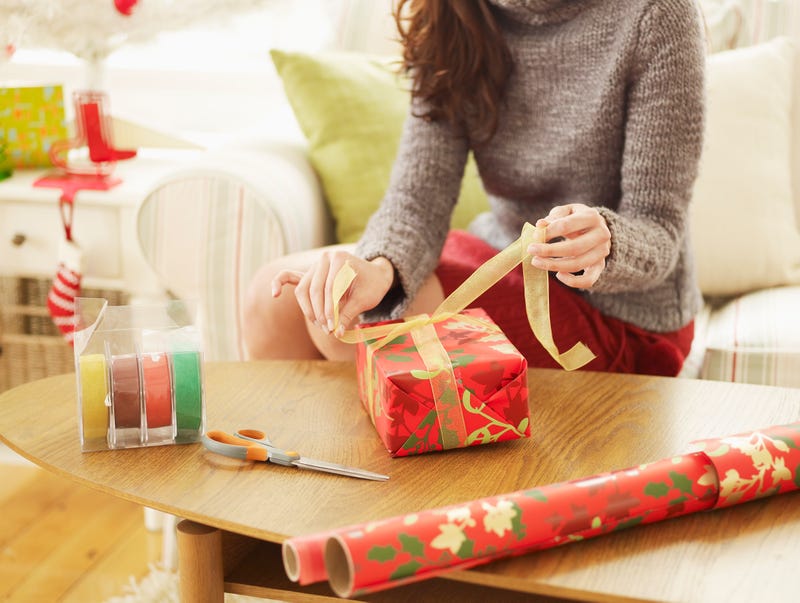 A woman ties a bow on a wrapped present with tape, scissors and a roll of wrapping paper lying on the table nearby