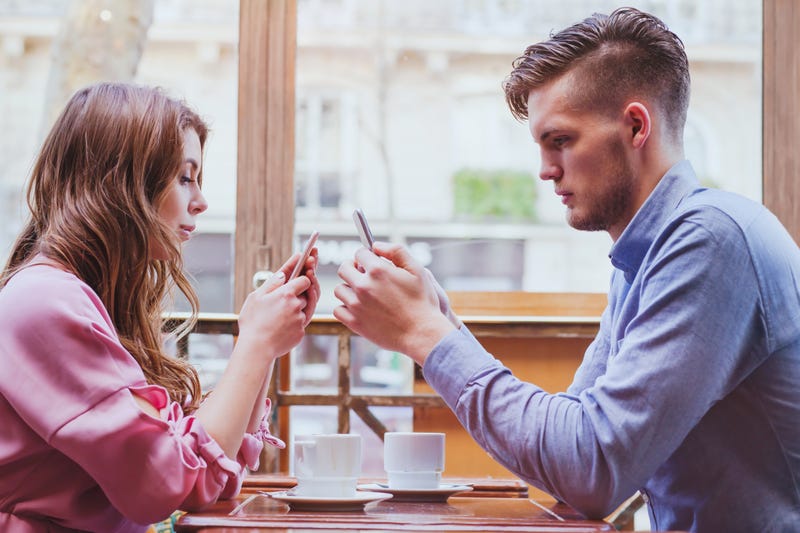 A couple sit across from each other at a cafe table both using their phones