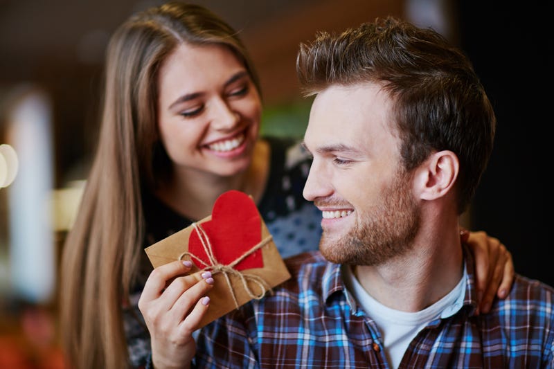 A woman leans over a man's shoulder to hand him a gift wrapped in brown paper with a large red paper heart affixed to it