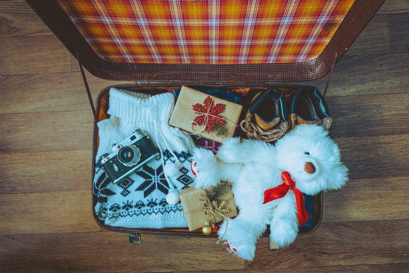 From above, an open suitcase loaded with a sweater, a stuffed bear, a camera, and wrapped gifts