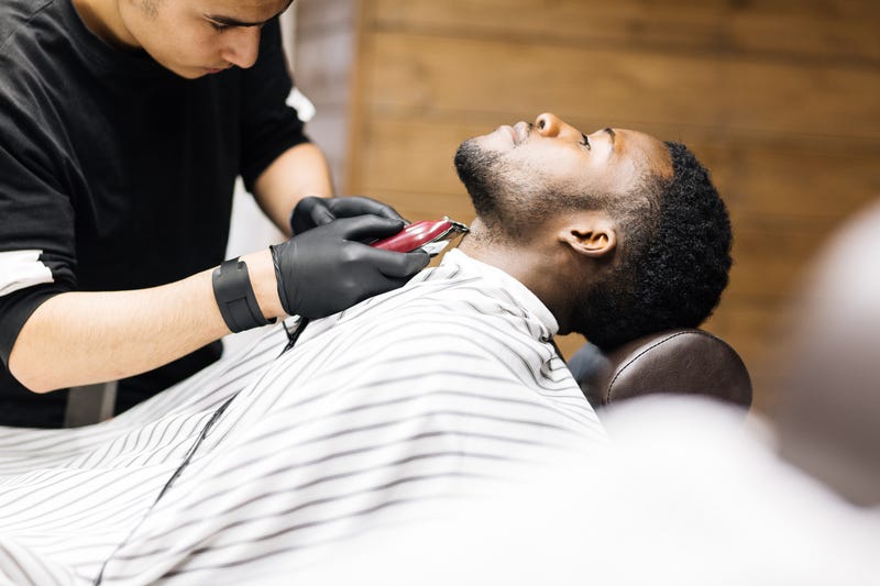 A barber trims a client's beard with an electric razor as he lies back in a chair with a smock