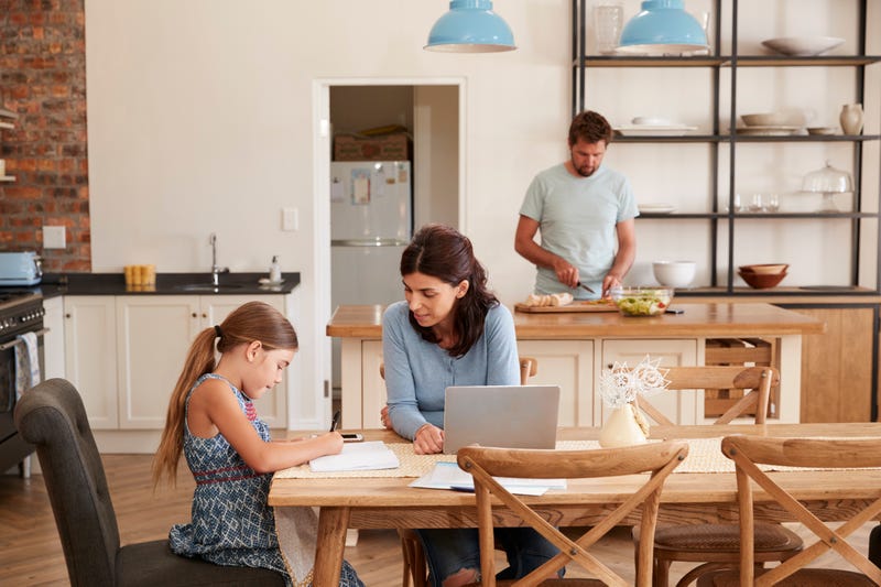 A mother helps her daughter with homework while a father works in the kitchen in the background