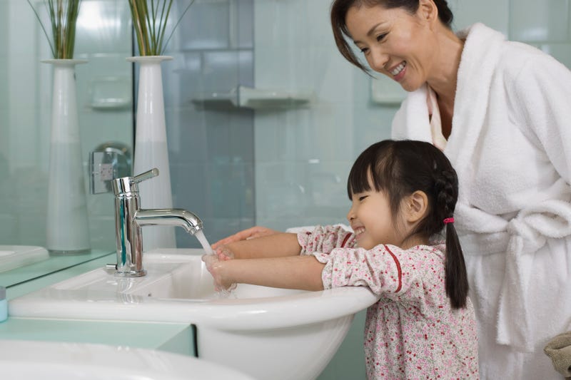 A mother stands over her daughter as she washes her hands in a sink