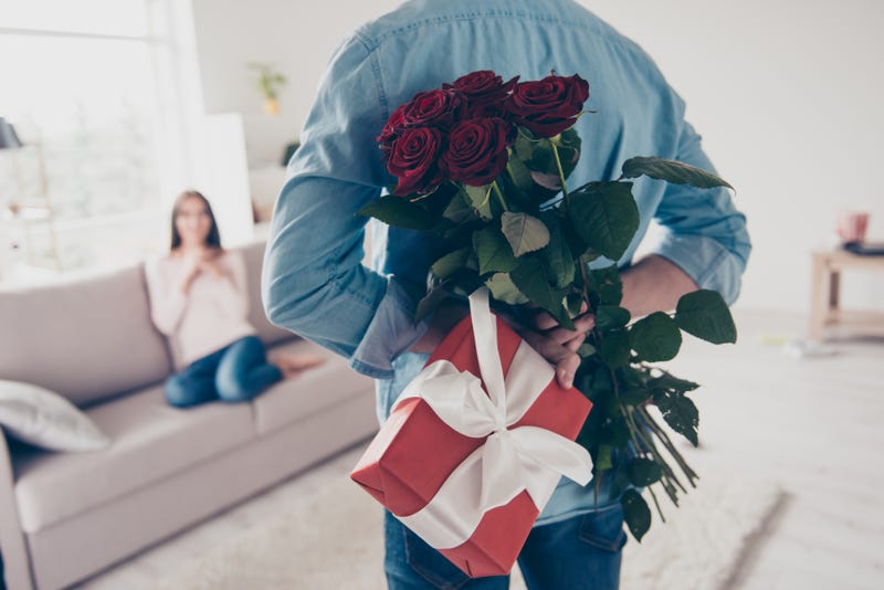 A man holds a wrapped gift and a bouquet of flowers behind his back while a woman waits on a couch in the background