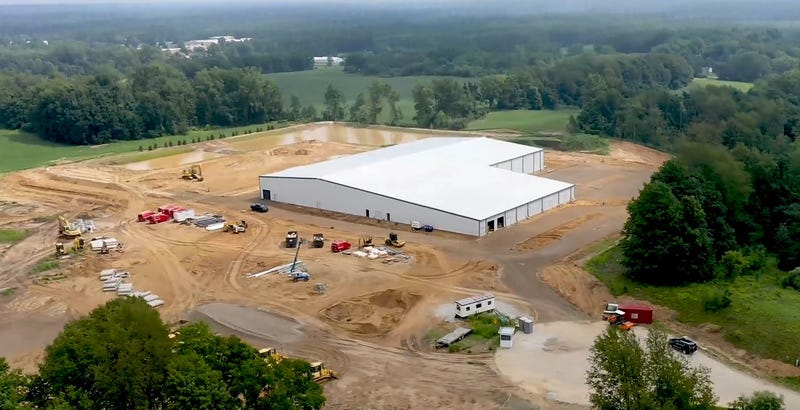 A large white building on a dirt lot surrounded by greenery and work trucks