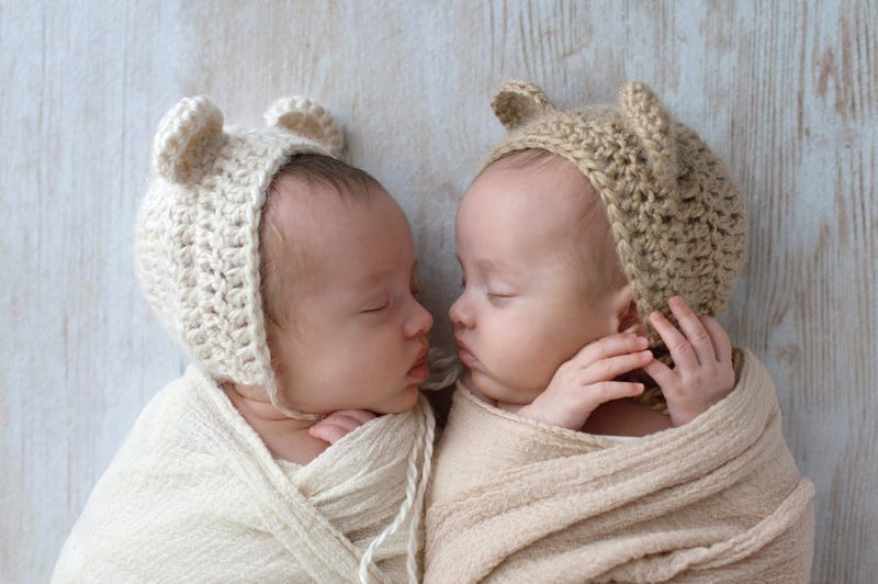 Profile headshot of two, fraternal, twin, baby girls sleeping. They are wearing crocheted bear hats and are swaddled in cream and tan wraps.