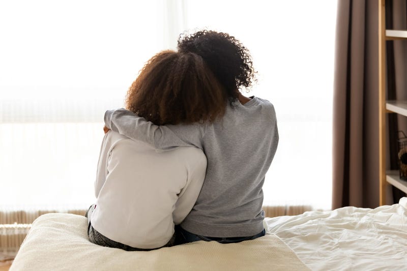 Two little girls, sitting on a bed, shown from behind, hugging each other.