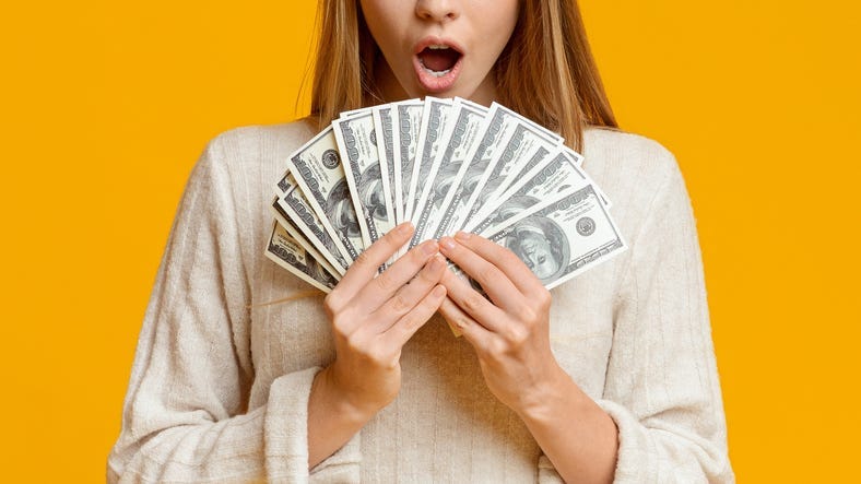Amazed young woman holding bunch of money banknotes, celebrating profit over orange background, panorama with copy space