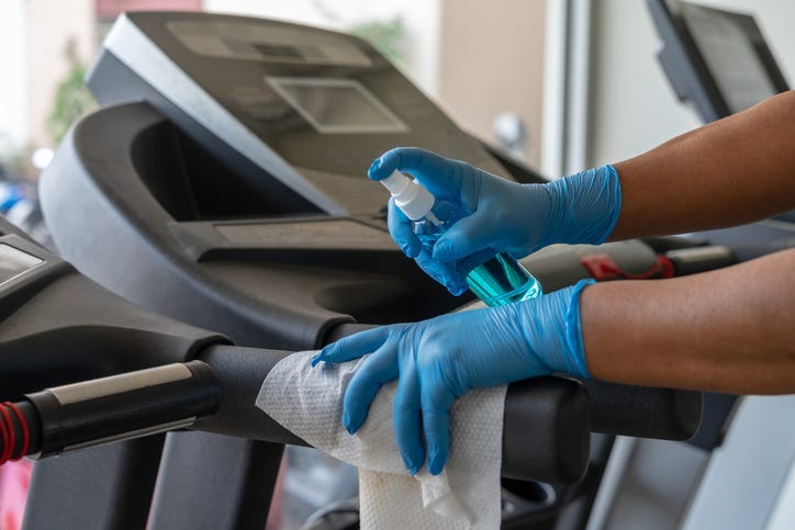 Staff using wet wipe and a blue sanitizer from the bottle to clean treadmill in gym
