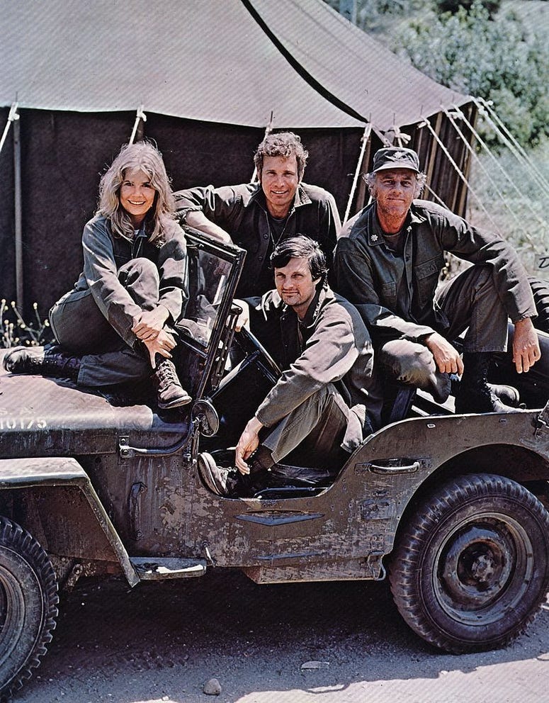 American actor, director and writer Alan Alda in the driving seat of a jeep, surrounded by Loretta Swit and other cast members of the hit television show M.A.S.H, in costume as members of a US Army medical corp.