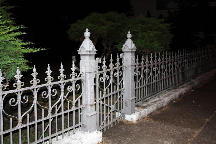 white stone or plaster fence with ornate wrought iron meeting at a creepy eerie cemetary gate with posts. taken in total darkness with one green tree on the side