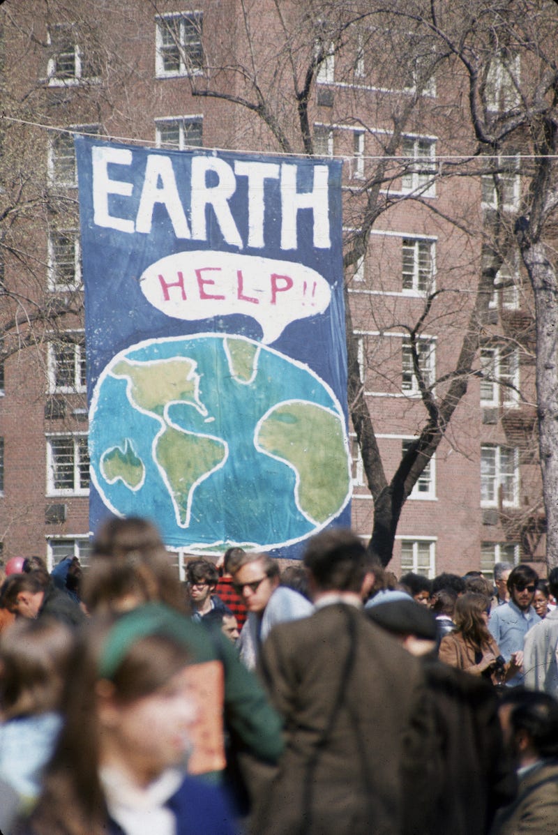 A crowd of people gather near a large poster that shows a speach bubble from planet Earth that reads 'Help!!', on the occaision of the first Earth Day conservation awareness celebration, New York, New York, April 22, 1970.