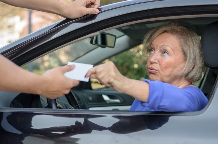 Concerned elderly woman driver handing over her licence to a man reaching towards the open window during a road block or after committing a minor offence