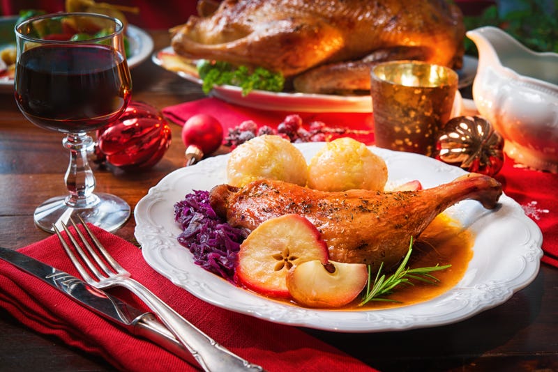 A turkey leg with fixins on a plate beside cutlery, a glass of wine, and Christmas ornaments