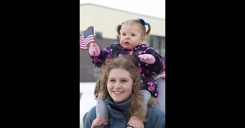 Brynlee, 1, sitting on top of family au pair, Jennifer Rauch, waves an American flag while waiting to greet her father at Joint Base Elmendorf-Richardson, Alaska, Feb. 7, 2020.