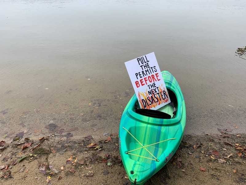 Kayak with sign that says "Pull the permits before the next disaster" at the shore of a lake.