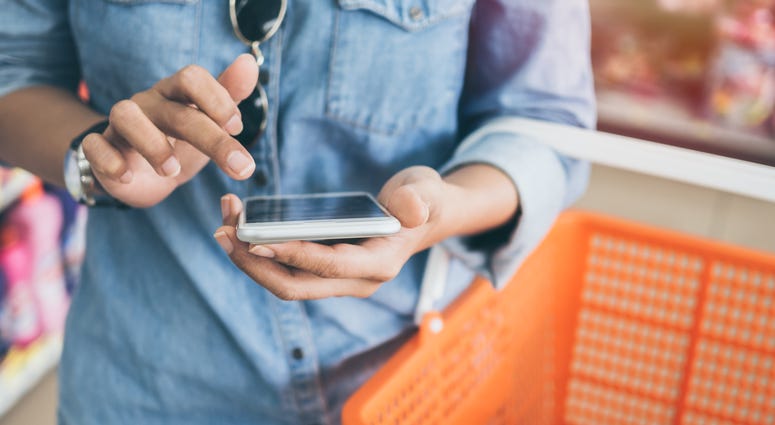 Woman wearing blue jeans shirt and sunglasses using mobile phone to compare price and holding orange shopping basket in mini mart background. Shopping basket - stock photo