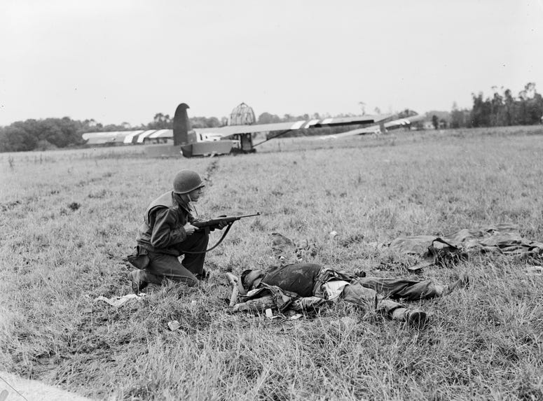1st July 1944: An American soldier crouching by the body of a dead German soldier with US Waco troop carrying gliders on the field behind him in Normandy.