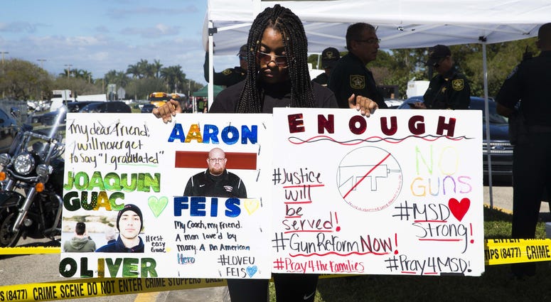 Tyra Hemats, 19, a 12th grader at Marjory Stoneman Douglas High School, stands with signs one advocating for gun reform and another honoring her coach and friend, both of whom were killed in Wednesday's shooting.