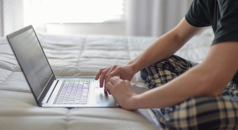 College student working on laptop computer on bed in morning light.